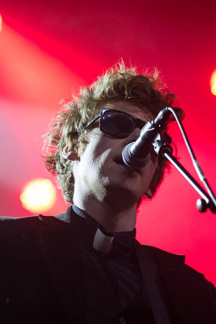 A musician singing into a microphone, wearing sunglasses and a black shirt with a clerical collar, playing guitar, with red stage lights in the background.