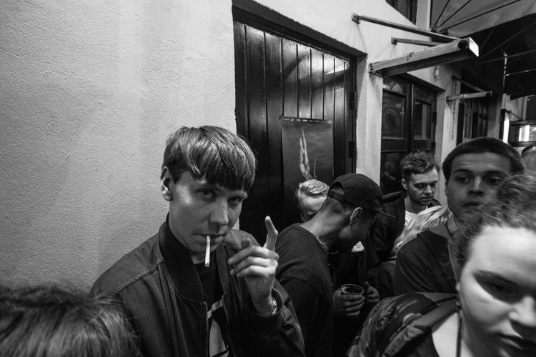 A group of people standing closely together at night outside a building, with one person in the foreground smoking a cigarette and flashing a peace sign.