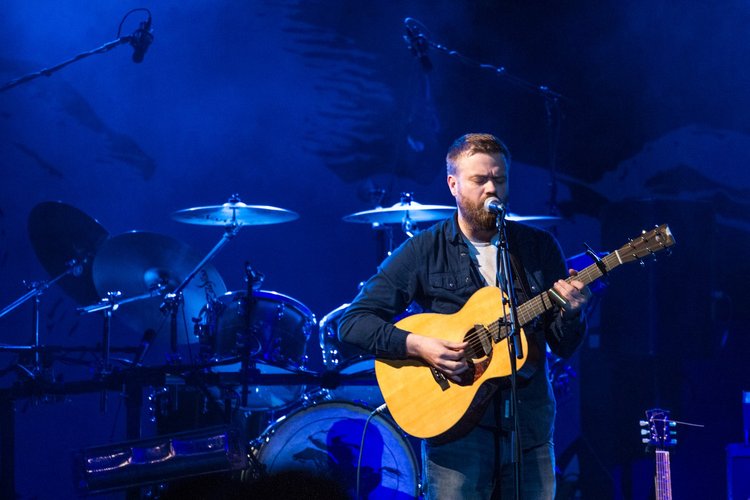 Man with a beard singing into a microphone while playing an acoustic guitar on stage, illuminated by blue lighting, with a drum set and a skeleton fish decoration in the background.