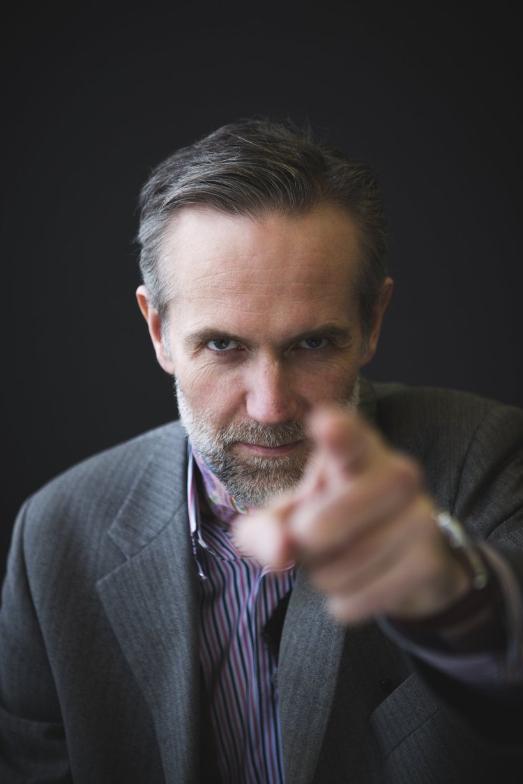 A man in a business suit pointing directly at the camera with a confident expression.
