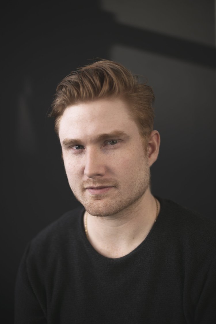 A young man with reddish hair and a light beard, wearing a black shirt, posing in front of a dark background.