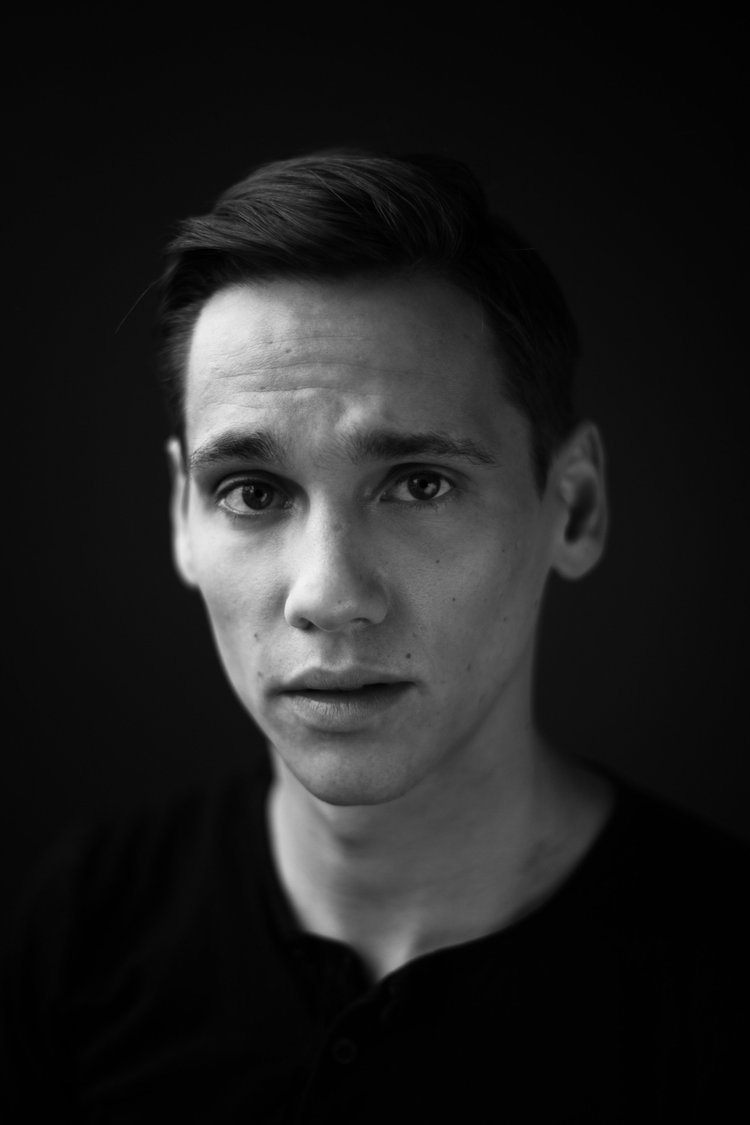 Black and white portrait of a young man with short hair, looking directly at the camera, with a dark background.