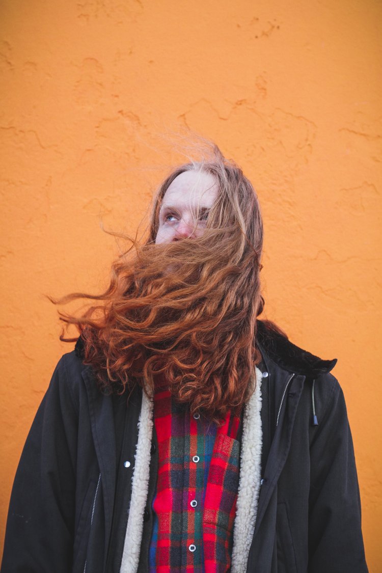 Young woman with long red hair and a plaid shirt standing against an orange wall.