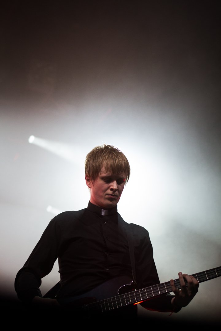 A young man playing an electric guitar on stage, wearing a black shirt and a clerical collar, with a dramatic backlight creating a silhouette effect.