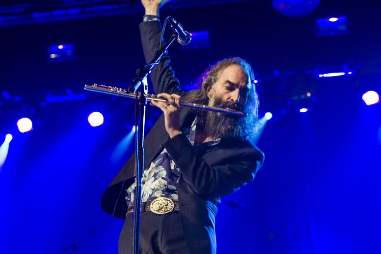 Man with long hair and beard playing flute on stage with blue lights.