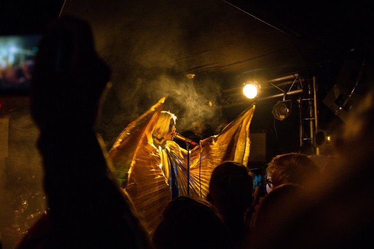 A performer on stage, dressed in a gold outfit with large gold wings, singing into a microphone under bright stage lights, with an audience in front.