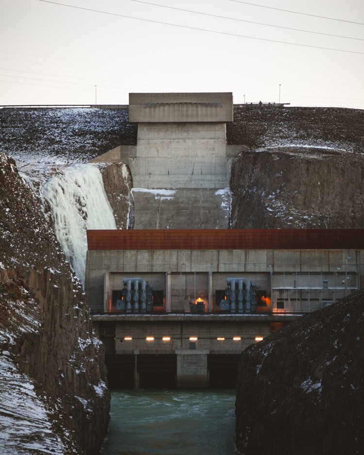 Hydroelectric dam with water spilling from the reservoir, concrete structure with power turbines below, snow on the surrounding rocky landscape.
