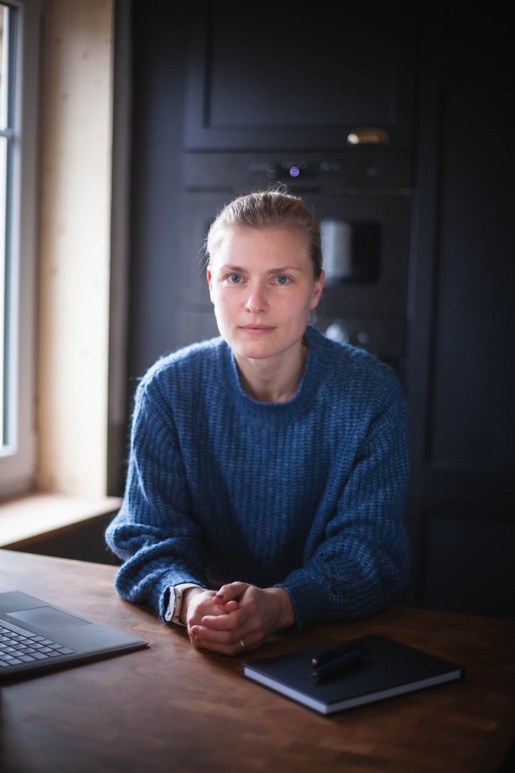 A woman with light hair and blue eyes, wearing a blue sweater, sitting at a wooden table in a room with dark cabinets and a window.