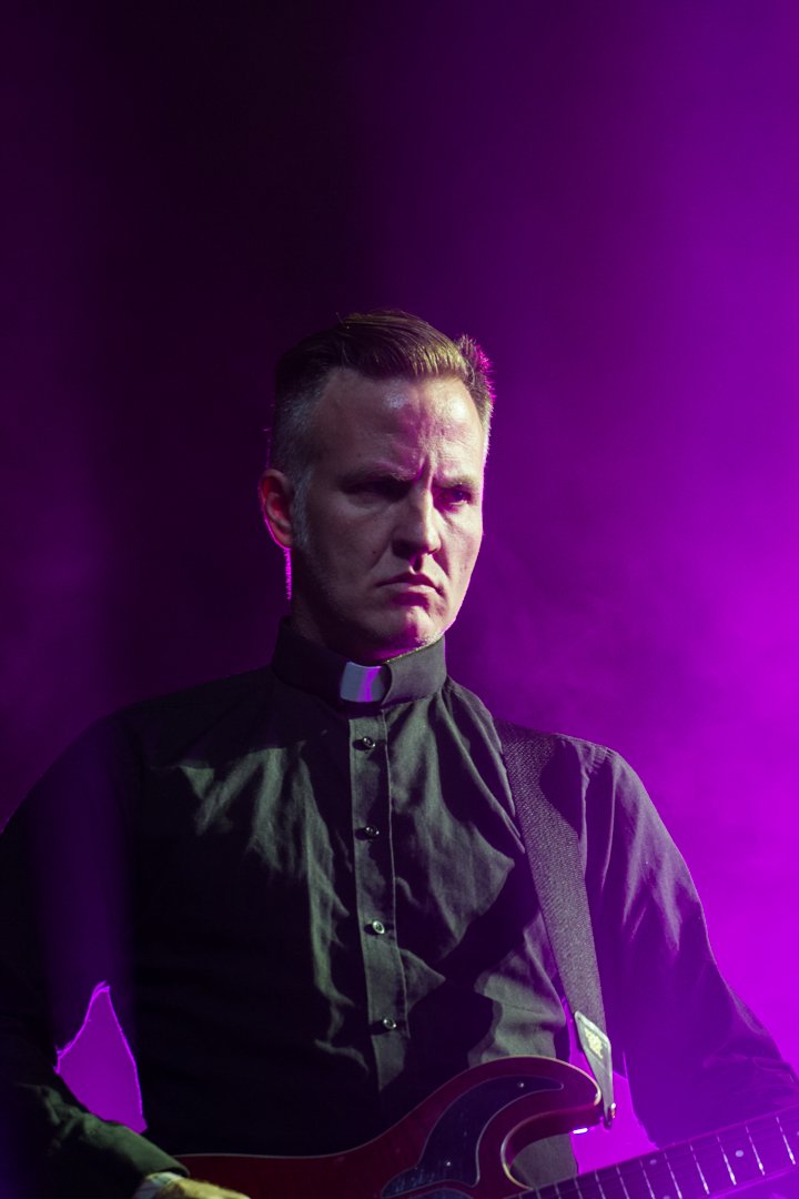A man dressed as a priest wearing a black clerical shirt with a white collar, playing an electric guitar under purple stage lighting.