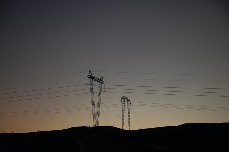Silhouette of power transmission towers and power lines against a dusk or dawn sky with a horizon line of rolling hills.