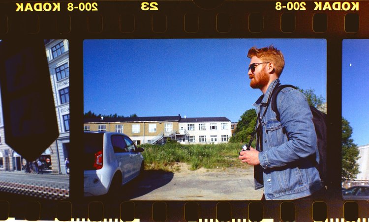 A young man with red hair, beard, sunglasses, wearing a denim jacket and backpack, walking outdoors on a sunny day with a clear blue sky, in an area with residential buildings, parked cars, and greenery.