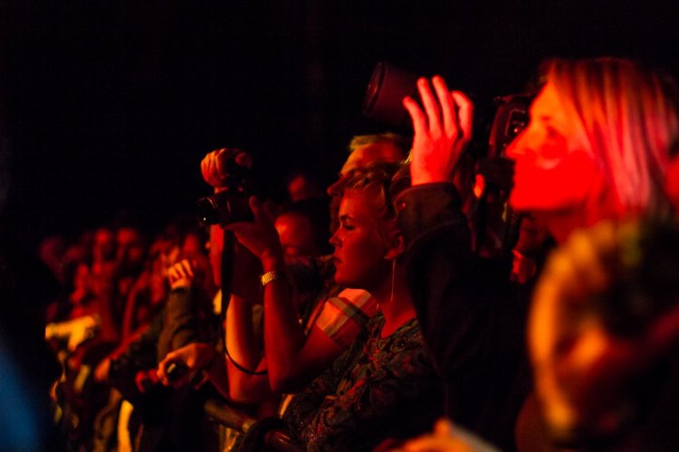 People sitting in a dark venue, some taking photos and videos, illuminated by red and orange lighting.