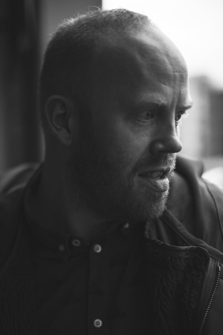 A close-up black and white portrait of a man with short hair and a beard, looking to the side, with a serious expression.