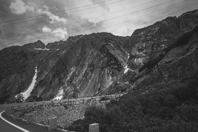 Black and white photo of a mountainous landscape with a road curving along the base of steep rocky cliffs. There are power lines across the sky, and some snow patches are visible on the mountain slopes.