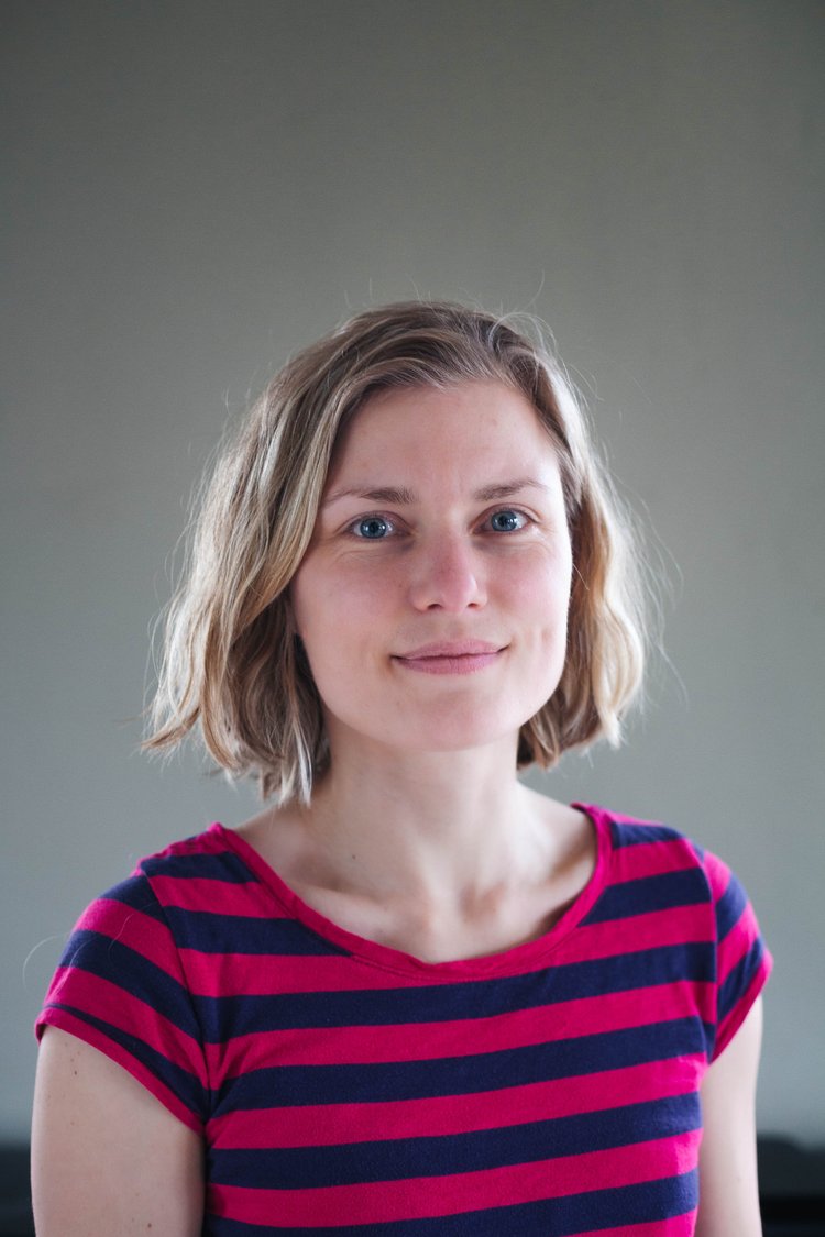 A woman with shoulder-length blonde hair and blue eyes, wearing a pink and navy striped t-shirt, smiling softly at the camera against a plain neutral background.