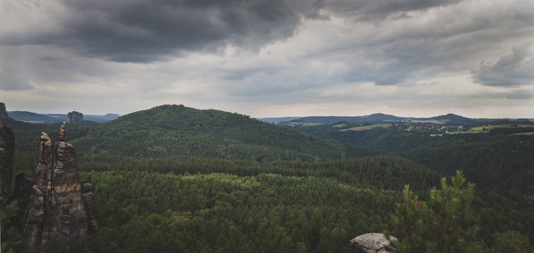 Landscape view of rolling hills covered in dense green forest with rock formations in foreground and a cloudy sky overhead.