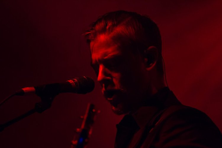A singer passionately performs with a microphone in a dark red-lit setting.