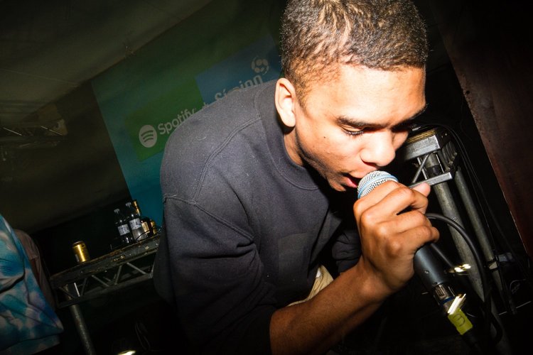 A young man speaking into a microphone during a performance, with a Spotify and SoundCloud banner in the background.