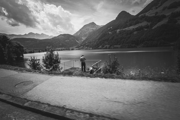 A person fishing by a lakeside with mountains in the background, black and white photograph.