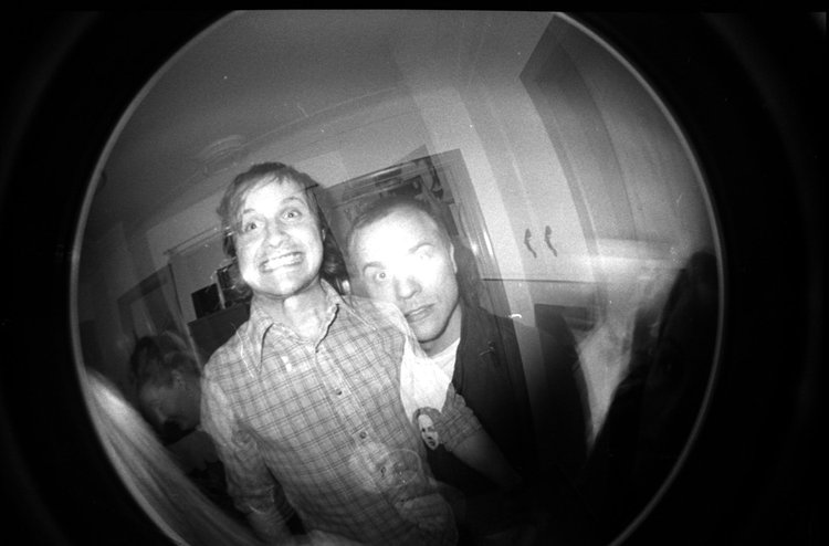 Two men with messy hair and casual clothes making funny faces for a photo taken with a fisheye lens, in an indoor setting.
