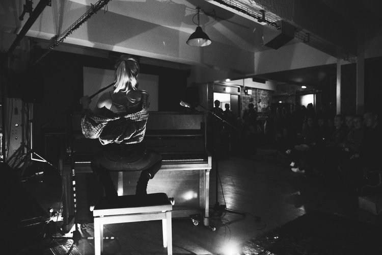 A woman with blonde hair performing on a piano in front of an audience in a dimly lit indoor venue.