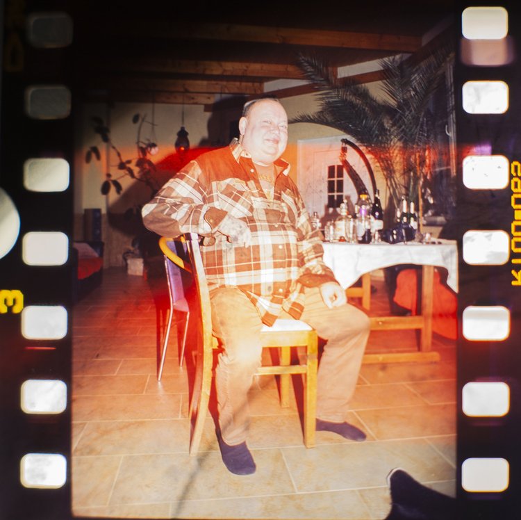 A man sitting on a wooden chair in a cozy indoor setting, with a table behind him holding bottles and glasses. The room has warm lighting, wooden ceiling panels, and decorative plants.