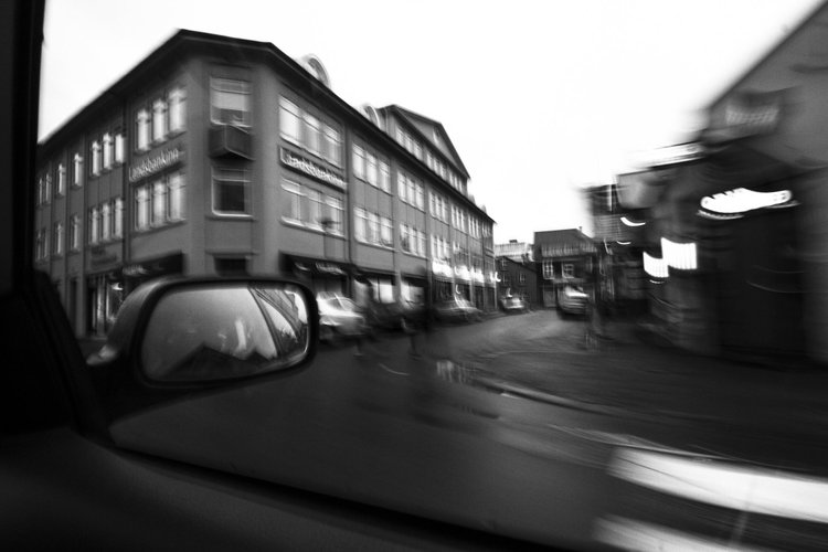 Black and white photo of a street scene with blurred buildings and cars viewed from inside a vehicle, with a reflection in the side mirror showing part of the vehicle's interior.