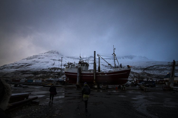 Fishing boat in a dockyard surrounded by snow-covered mountains under a dark, cloudy sky.