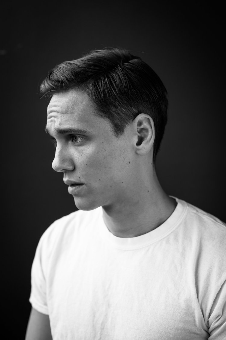 Black and white portrait of a young man in profile with a short haircut, wearing a white t-shirt, against a dark background.