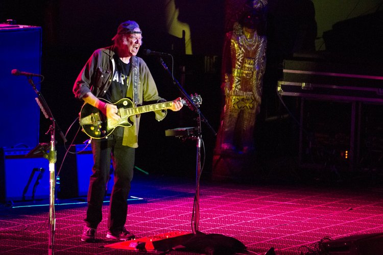 An older man with a backward cap playing an electric guitar on stage with colorful lighting.