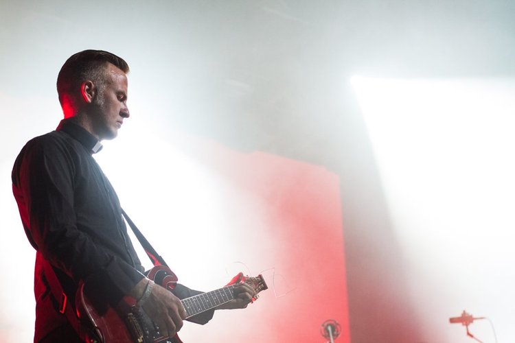 A male musician wearing a black shirt playing an electric guitar on stage, illuminated by bright lighting.