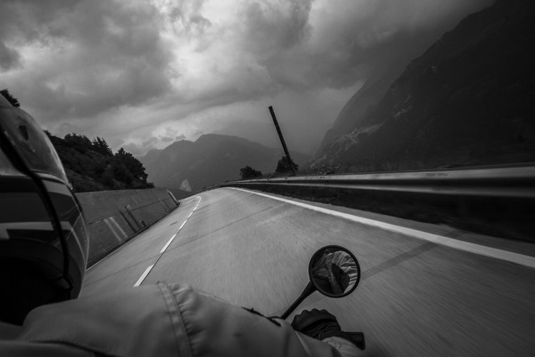 Black and white photo of a motorcycle rider on a winding mountain road with dark clouds in the sky and mountain scenery in the background.