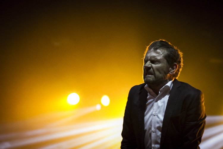 A man in a suit with a white shirt, grimacing with his eyes closed, standing in a dark space illuminated by bright yellow stage lights in the background.