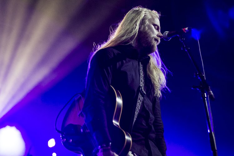 A male musician with long blonde hair and a beard performing on stage with a microphone and guitar, illuminated by purple and blue stage lighting.
