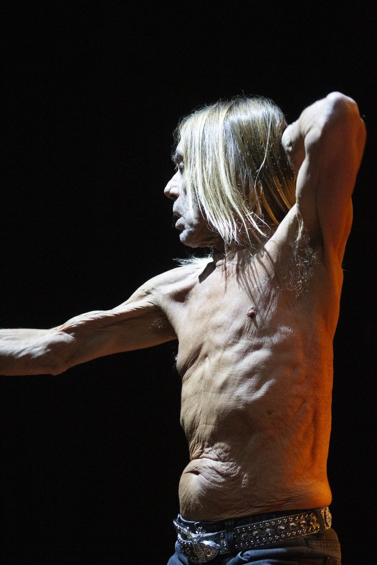 A topless man with long blonde hair stretching his arm behind his head, standing in profile against a dark background.