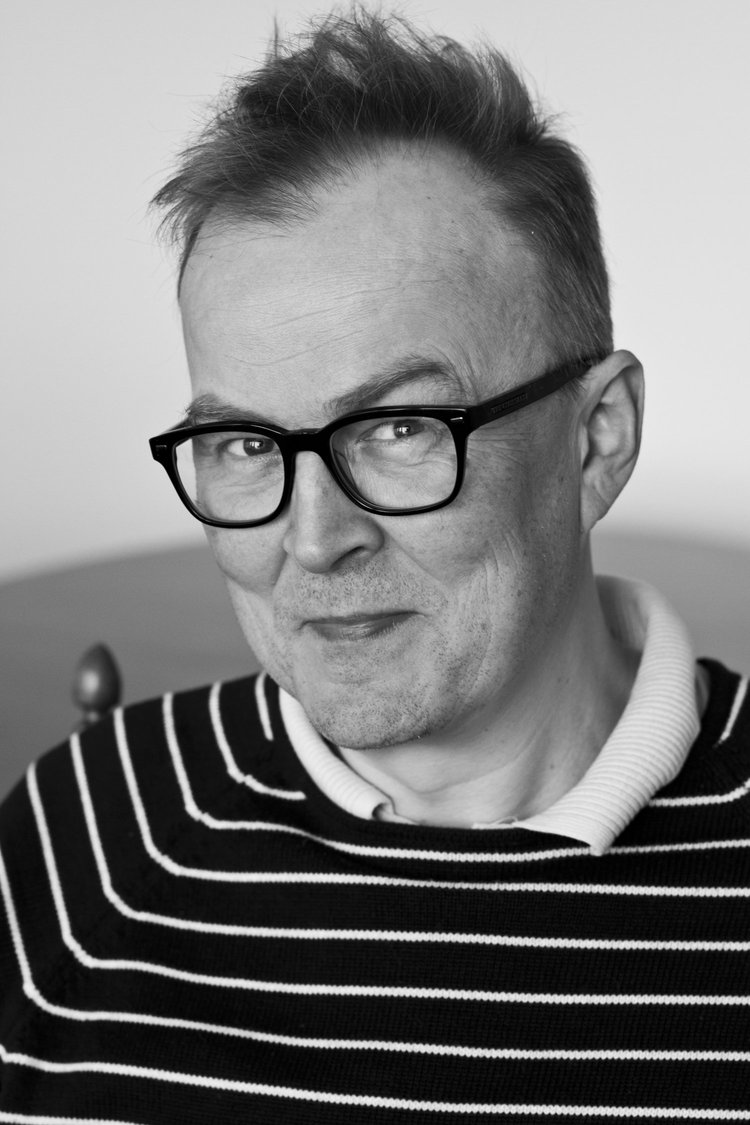 A black-and-white portrait of a man wearing glasses and a striped sweater, smiling softly.