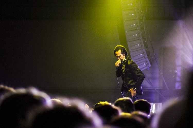Male performer dressed in black with yellow accents singing into a microphone on stage with yellow and purple lighting, and a large speaker system in the background, in front of an audience.