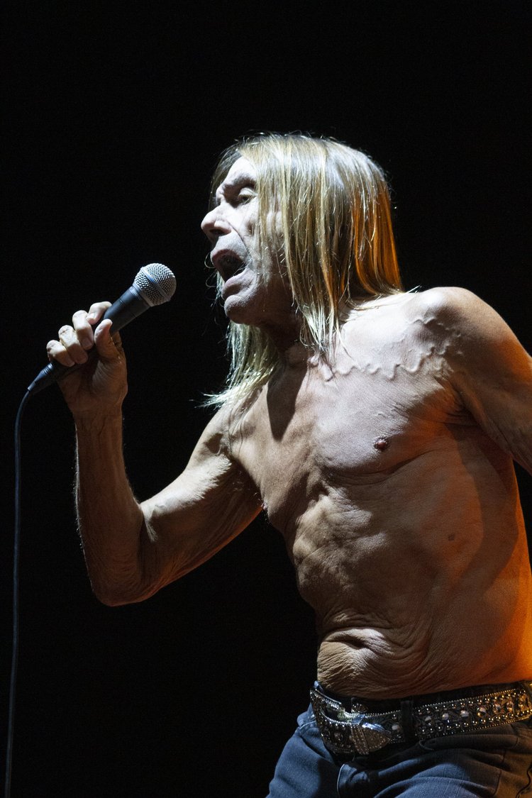 A shirtless man with long blonde hair singing into a microphone on a dark stage.