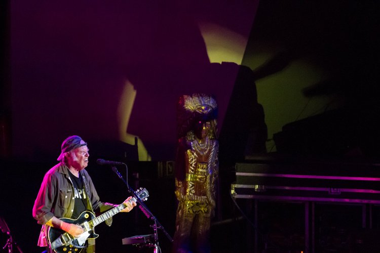 A man is playing an electric guitar and singing into a microphone on stage, wearing a cap, glasses, a black t-shirt, and an olive jacket. There is colorful stage lighting and a dark background.