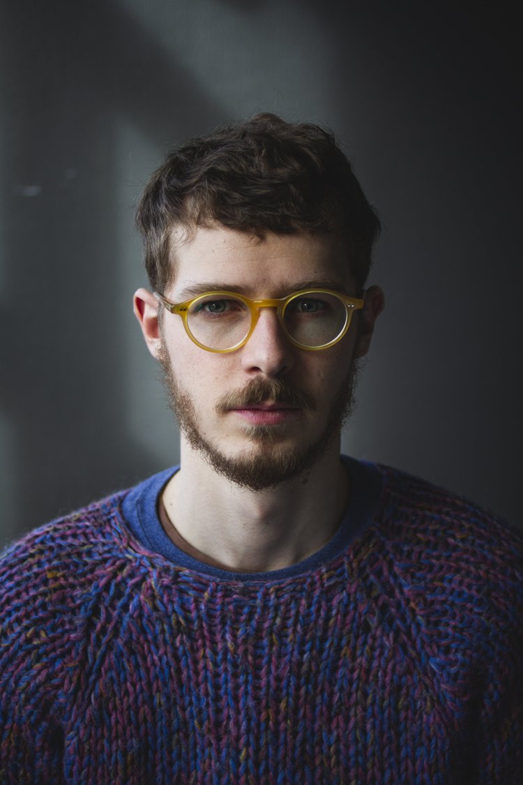 A young man with light skin, brown hair, and a beard wearing yellow glasses and a colorful knitted sweater against a dark background.