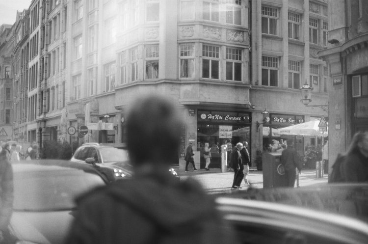 Street scene with pedestrians walking past a café named Hana Cuisine and Hana Bar on a corner building. Cars parked along the street, and a person with blurred head in foreground. Black and white photo.