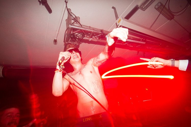 A shirtless man with tattoos wearing sunglasses, holding a microphone, and singing. A person next to him is pointing with a finger. The scene is illuminated with red lighting and there is stage equipment overhead.