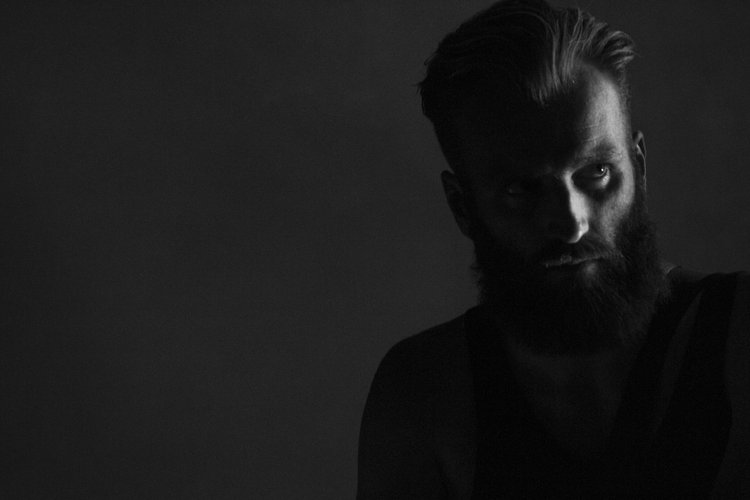Black and white photo of a man with a beard and styled hair, looking intensely at the camera in dim lighting.