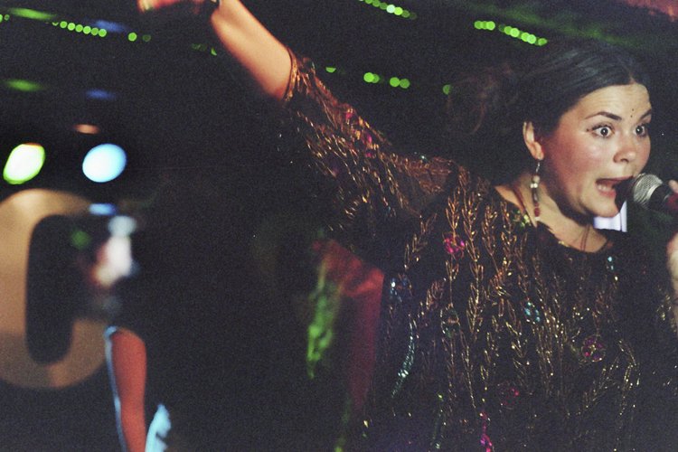 A woman singing into a microphone with her arm raised, wearing a colorful, patterned dress and earrings, on a stage with green and blue lights.