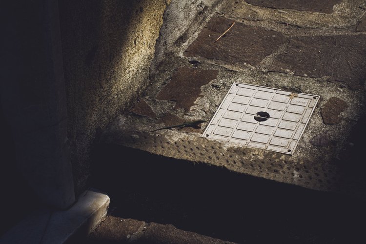 A street drain cover embedded in a cobblestone sidewalk, with surrounding stones and concrete.