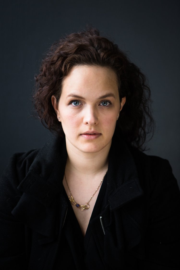 Portrait of a woman with curly brown hair, blue eyes, and light skin, wearing a black jacket and gold necklace, against a dark gray background.
