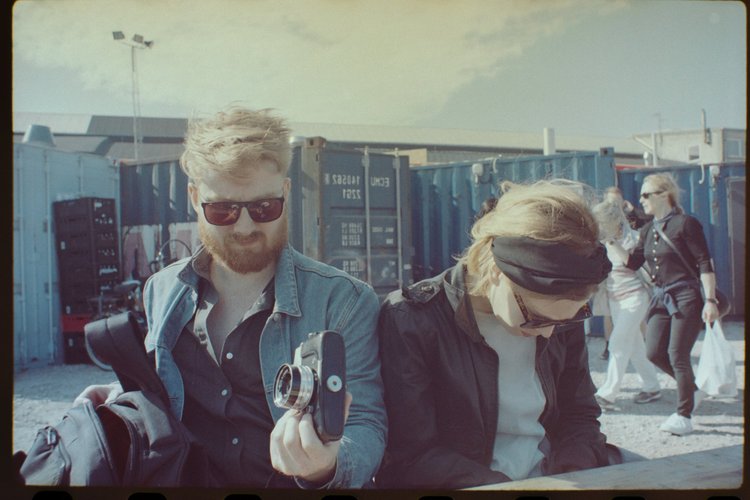 Two individuals sitting outdoors near blue shipping containers, with several other people walking or standing in the background. One person with sunglasses and a denim jacket is holding a camera, the other with a headband and glasses appears to be lo