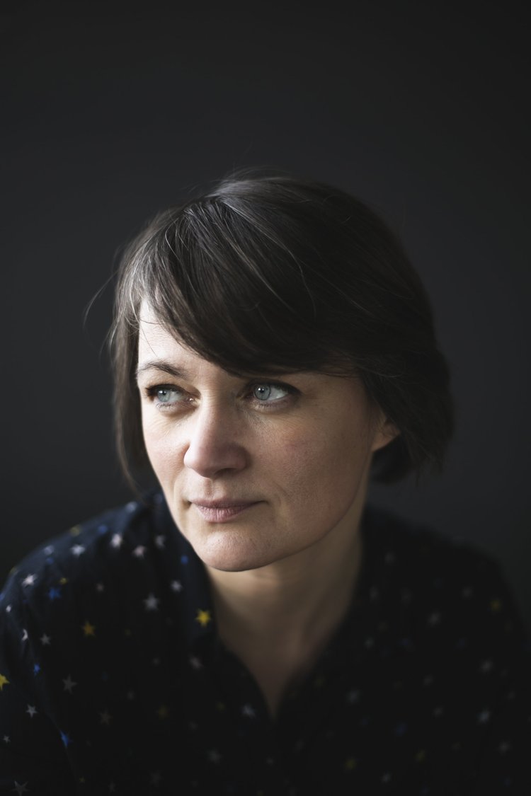 Close-up of a woman with short dark hair looking to the side, wearing a dark top with star patterns, against a dark blurred background.