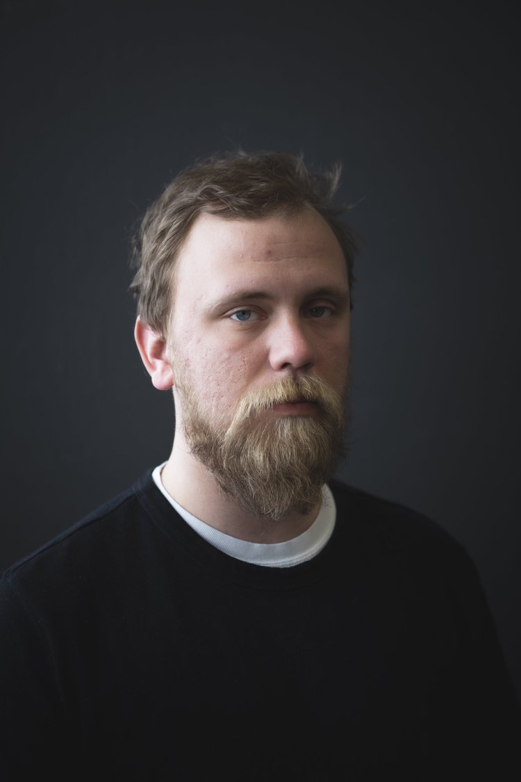 A portrait of a man with a beard and mustache, wearing a black shirt with a white collar, against a dark background.