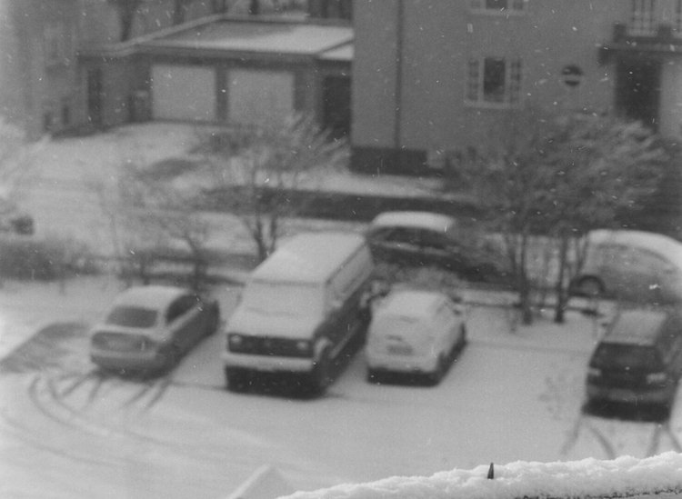 Snow-covered parking lot with several parked cars, snow-laden trees, and residential buildings in the background, taken during snowy weather.